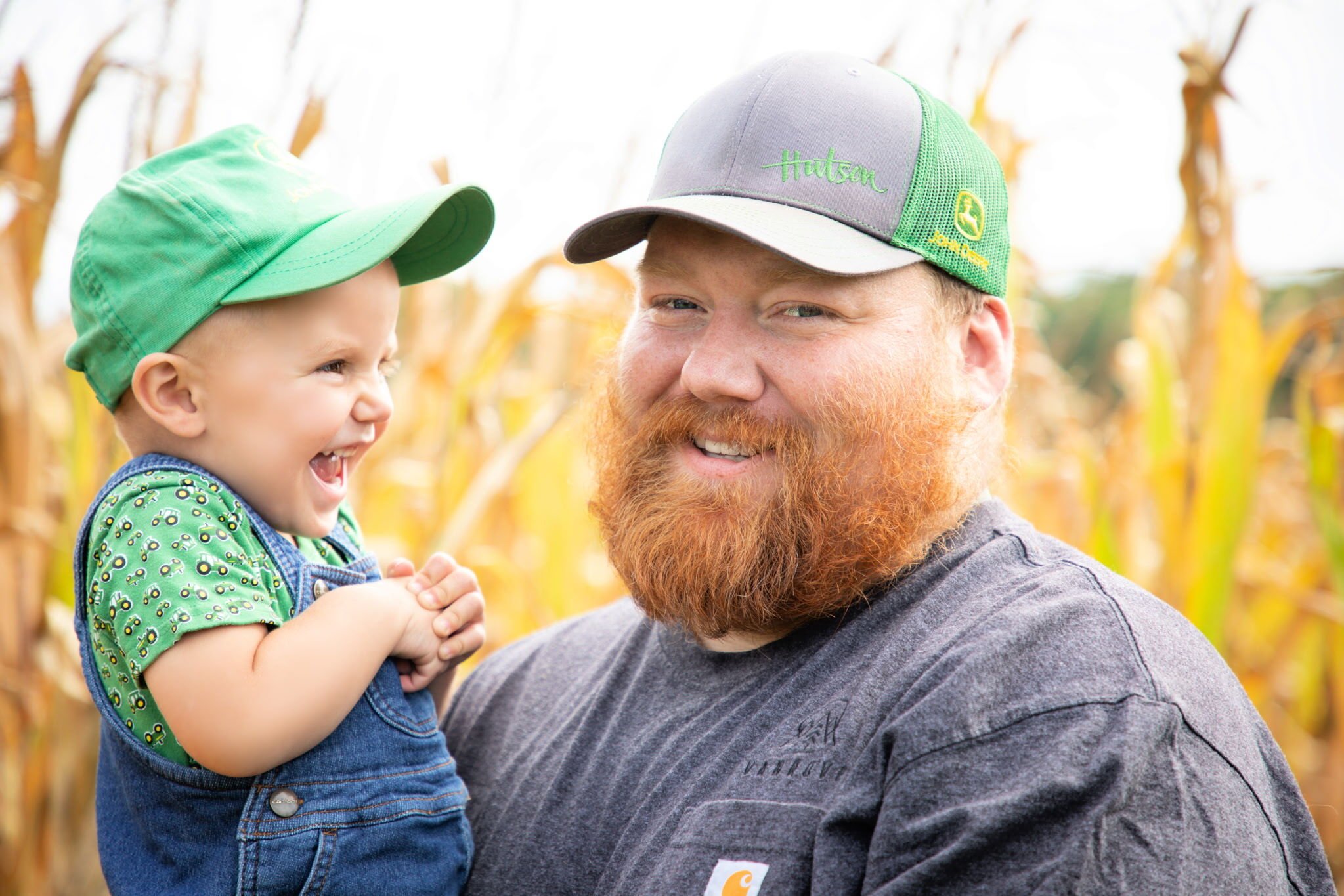 bearded man holding his young son, both are smiling, corn field in the background